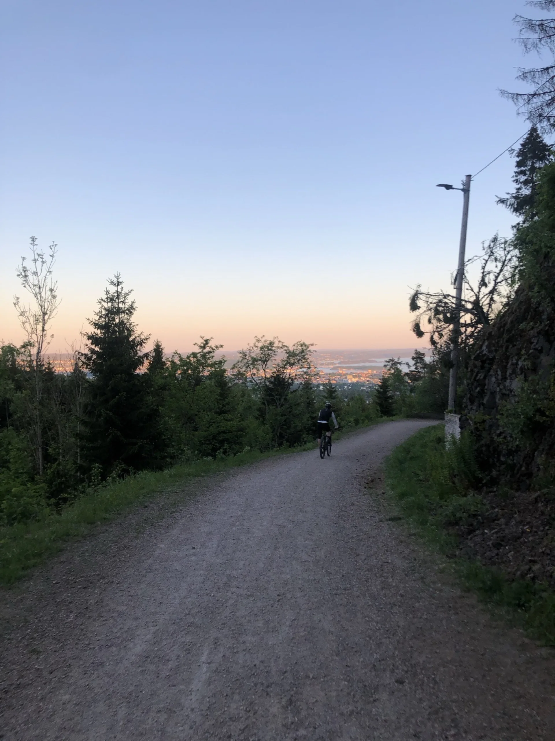 A rider on a Nordmarka gravel road at dusk, with Oslo and the fjord lit by the setting sun below