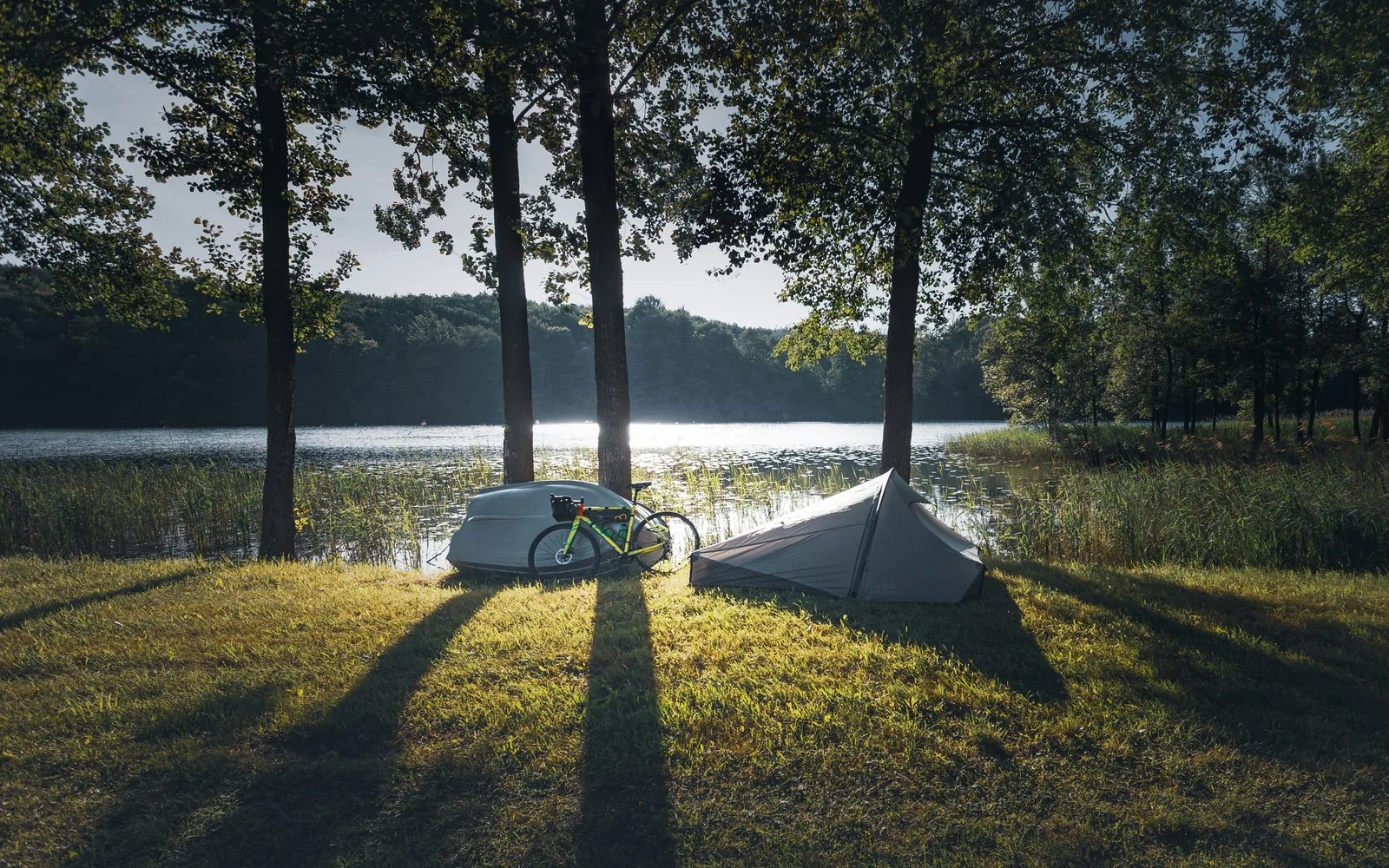 A bikepacking bike and a tent pitched beside a forest lake at golden hour
